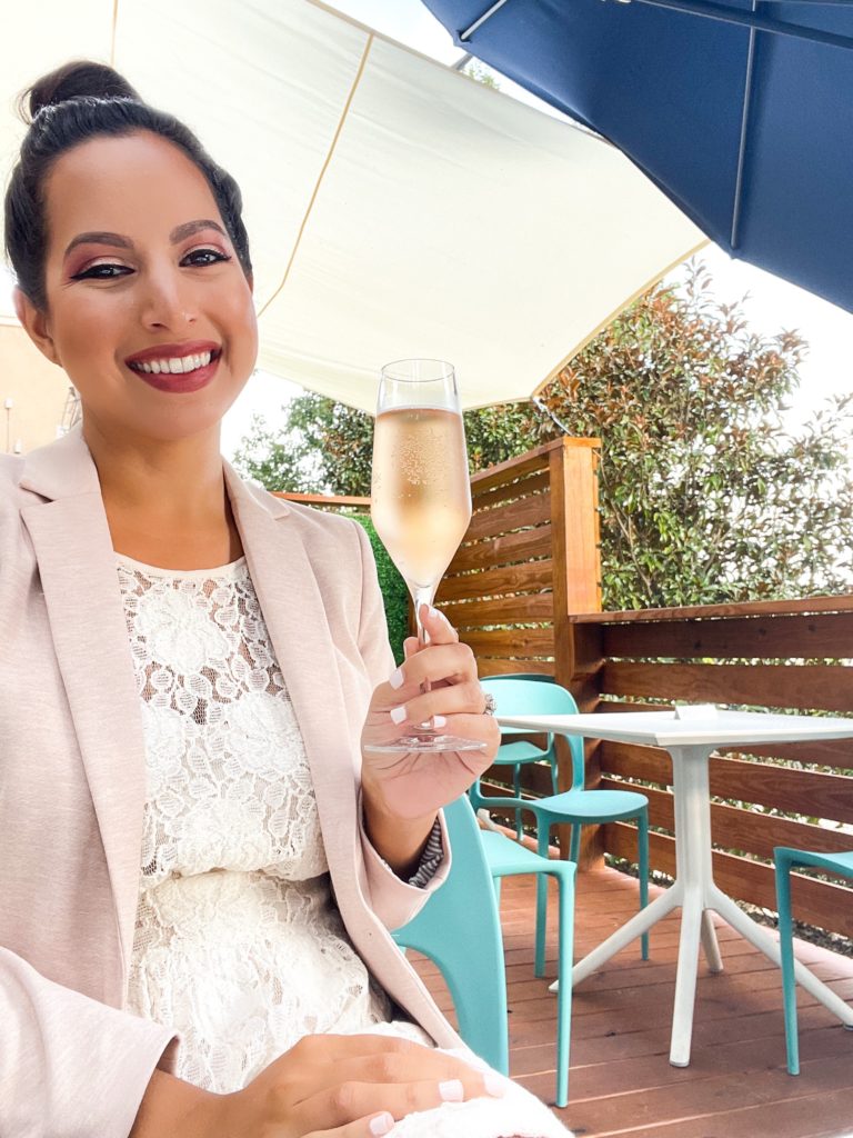 Maria Hossain - owner, smiling and holding a glass of champagne, while seated outdoors on a deck, wearing a white dress and pale pink blazer. 